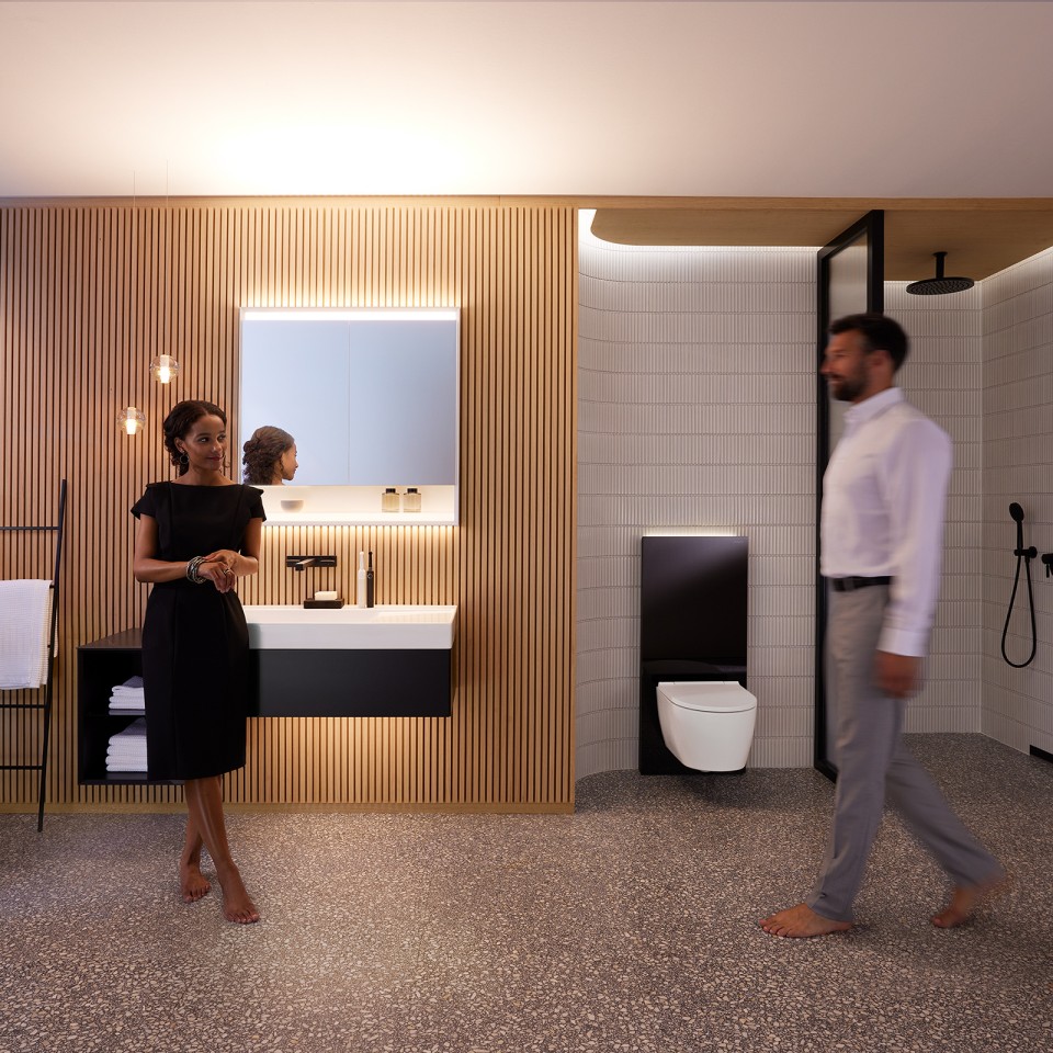 Homem e mulher numa casa de banho perfeitamente iluminada com móveis de casa de banho e sanitários Geberit ONE (© Geberit)
